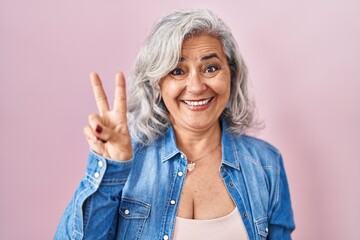 Middle age woman with grey hair standing over pink background showing and pointing up with fingers number two while smiling confident and happy.