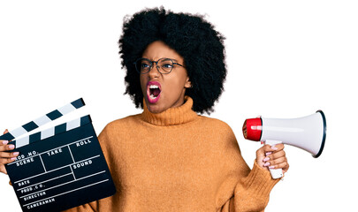 Young african american woman holding video film clapboard and megaphone angry and mad screaming...