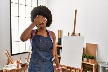 Young african american woman with afro hair at art studio covering eyes with hand, looking serious...