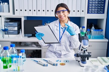 Hispanic young woman working at scientist laboratory cheerful with a smile on face pointing with hand and finger up to the side with happy and natural expression