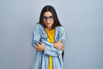 Young hispanic woman standing over blue background shaking and freezing for winter cold with sad and shock expression on face