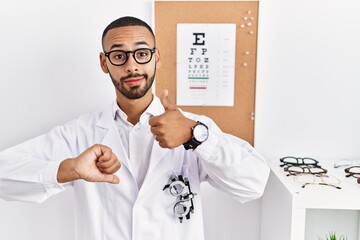 African american optician man standing by eyesight test doing thumbs up and down, disagreement and agreement expression. crazy conflict