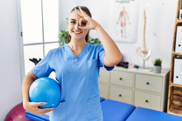 Young physiotherapist woman holding pilates ball at medical clinic smiling happy doing ok sign with...