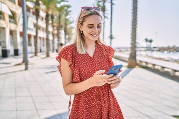 Young blonde girl smiling happy using smartphone at the promenade.