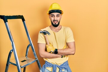Handsome man with beard wearing hardhat holding hammer by stairs clueless and confused expression....
