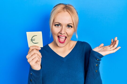 Young blonde woman holding euro symbol reminder celebrating achievement with happy smile and winner expression with raised hand