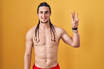 Fototapeta premium Hispanic man with long hair standing shirtless over yellow background showing and pointing up with fingers number three while smiling confident and happy.