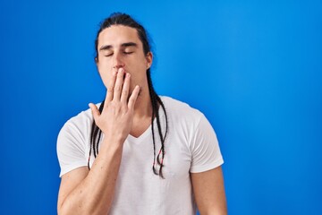 Hispanic man with long hair standing over blue background bored yawning tired covering mouth with hand. restless and sleepiness.