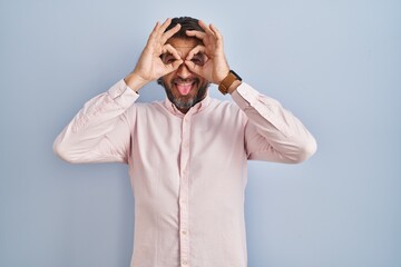 Handsome middle age man wearing elegant shirt background doing ok gesture like binoculars sticking tongue out, eyes looking through fingers. crazy expression.