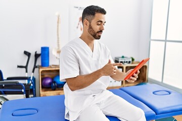 Young hispanic man wearing physiotherapist uniform having video call at clinic