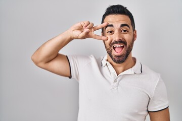 Young hispanic man with beard wearing casual clothes over white background doing peace symbol with fingers over face, smiling cheerful showing victory