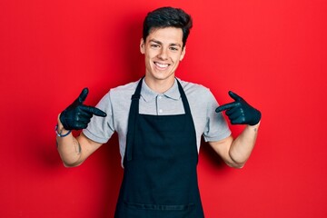 Young hispanic man wearing apron looking confident with smile on face, pointing oneself with fingers proud and happy.