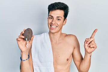 Young hispanic man naked holding pumice stone smiling happy pointing with hand and finger to the side - obrazy, fototapety, plakaty