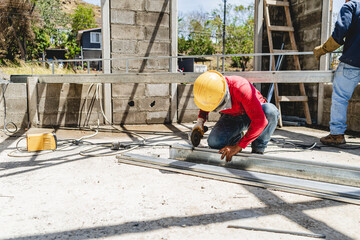 Image of an unknown man in safety gear, crouched down cutting a metal pipe at a construction site with a tool. 