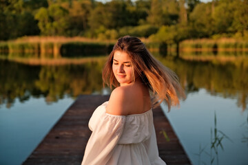 a beautiful girl is standing on the pier. in the background there is a river, a lake.