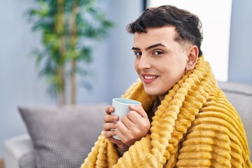 Young non binary man drinking coffee sitting on sofa at home