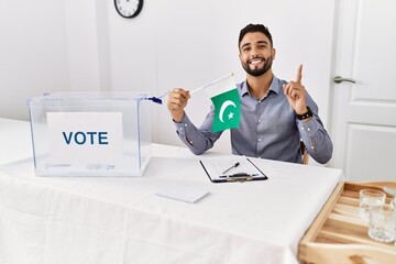 Young handsome man with beard at political campaign election holding pakistan flag surprised with an idea or question pointing finger with happy face, number one - obrazy, fototapety, plakaty