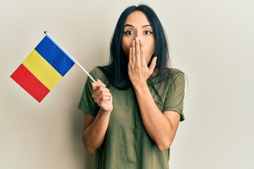 Young hispanic girl holding romania flag covering mouth with hand, shocked and afraid for mistake....