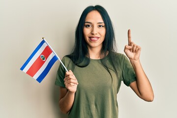 Young hispanic girl holding costa rica flag smiling with an idea or question pointing finger with...
