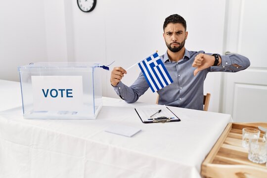 Young handsome man with beard at political campaign election holding greece flag with angry face, negative sign showing dislike with thumbs down, rejection concept