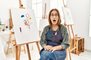 Middle age artist woman at art studio afraid and shocked with surprise expression, fear and excited...