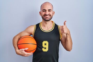 Young bald man with beard wearing basketball uniform holding ball doing happy thumbs up gesture with hand. approving expression looking at the camera showing success.