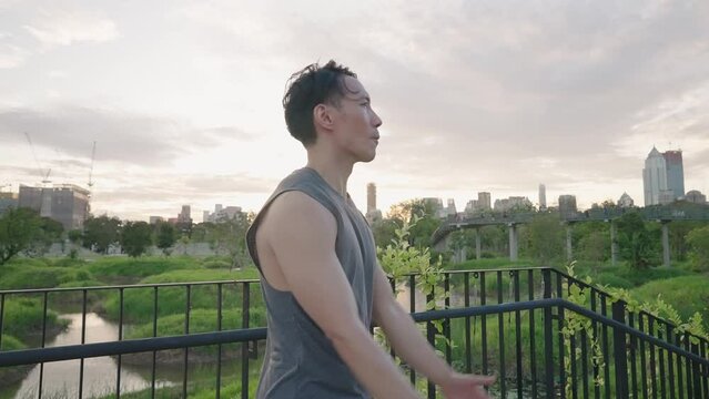 Asian Male Athlete Stretching Neck Shoulder Arms and Back Muscles against city sunset inside natural park. Active handsome young adult doing exercises outdoors at dawn. Urban Lifestyle. Health Care