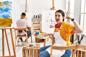 Young artist woman painting on canvas at art studio smiling amazed and surprised and pointing up with fingers and raised arms.