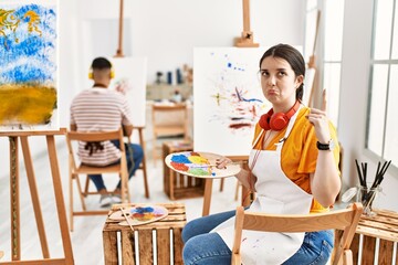 Young artist woman painting on canvas at art studio pointing up looking sad and upset, indicating...