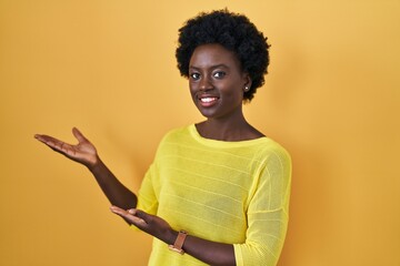 African young woman standing over yellow studio inviting to enter smiling natural with open hand