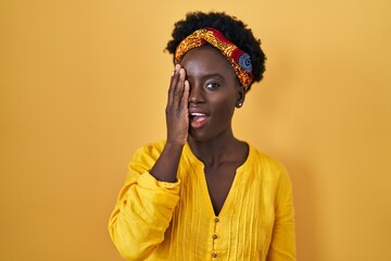 African young woman wearing african turban covering one eye with hand, confident smile on face and surprise emotion.