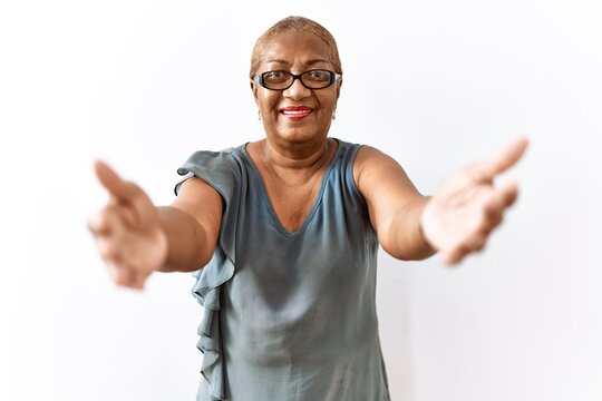 Mature hispanic woman wearing glasses standing over isolated background looking at the camera smiling with open arms for hug. cheerful expression embracing happiness.