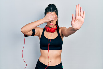 Young hispanic girl wearing gym clothes and using headphones covering eyes with hands and doing...