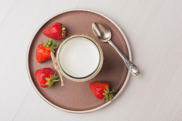 Top view on ceramic plate with homemade Greek yogurt in a glass jar and strawberry on wooden table. Calcium dairy fresh product.