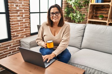 Middle age hispanic woman using laptop and drinking coffee at home