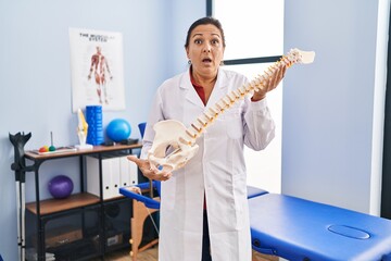 Middle age hispanic woman holding anatomical model of spinal column in shock face, looking...