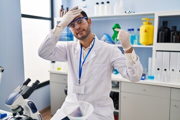 Young hispanic man with beard working at scientist laboratory holding green ribbon stressed and frustrated with hand on head, surprised and angry face