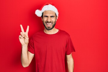 Young hispanic man wearing christmas hat smiling with happy face winking at the camera doing victory sign. number two.