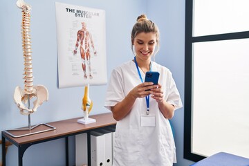 Young blonde woman wearing physiotherapist uniform using smartphone at clinic - obrazy, fototapety, plakaty
