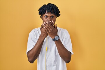 Young african man with dreadlocks standing over yellow background shocked covering mouth with hands...
