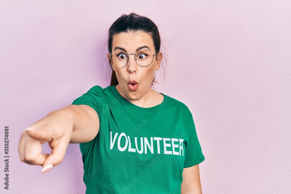 Sticker young hispanic woman wearing volunteer t shirt pointing with finger surprised ahead, open mouth amaz