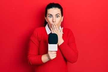Young hispanic woman holding reporter microphone covering mouth with hand, shocked and afraid for...