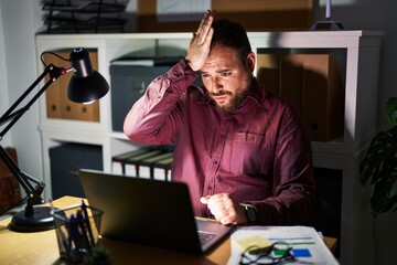 Plus size hispanic man with beard working at the office at night surprised with hand on head for...