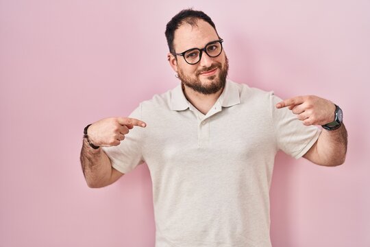 Plus size hispanic man with beard standing over pink background looking confident with smile on face, pointing oneself with fingers proud and happy.