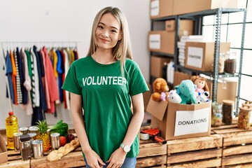 Asian young woman wearing volunteer t shirt at donations stand looking confident at the camera smiling with crossed arms and hand raised on chin. thinking positive.