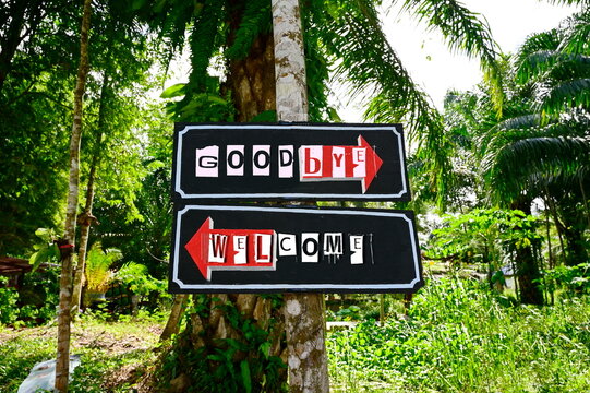 A wooden sign painted in black with red arrow pointing to the right written in white "Goodbye" and an arrow pointing to the left saying "Welcome". Two wooden signs mounted on tree at entrance the park