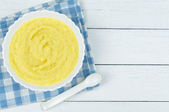 Corn porridge for a baby made of ground cereals in a white bowl with a banana on a blue napkin on a white wooden background. Space for text. Baby's first complementary food, baby nutrition.