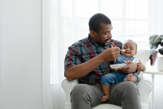 African toddler baby girl crying and don't want to eat food from her father who feeding with spoon while sitting on sofa at home. Little child care and relationship of dad and little daughter concept