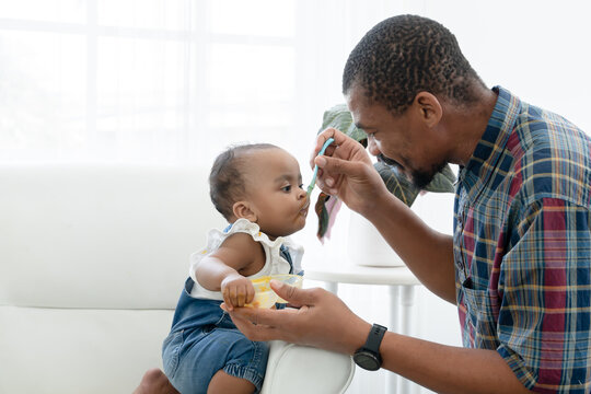 African father feeding adorable toddler baby girl with spoon while sitting on sofa at home and kid's face mess up with food. Little child care and relationship of dad and little daughter concept