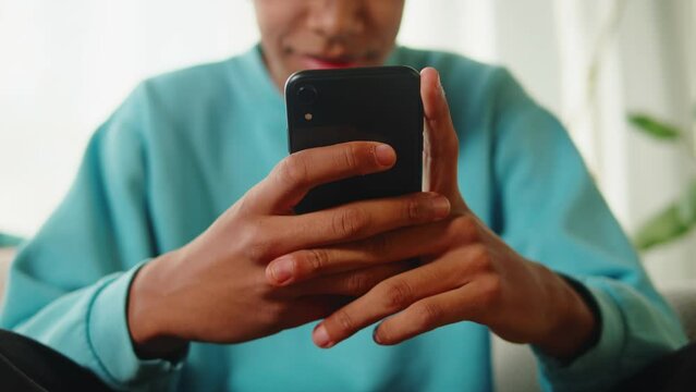 African American man using smartphone. Young guy texting at phone, teenager relaxing at home. Communicating with family and friends online, sitting on sofa in living room.